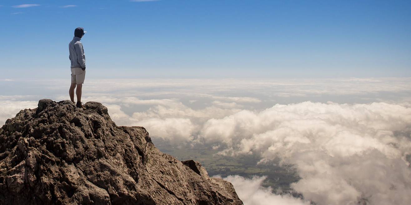 Sinnbild für eine klare, eindeutige Positionierung eines Unternehmens am Markt und für eine deutliche Abgrenzung vom Wettbewerb. Das Bild zeigt einen einzelnen Menschen auf dem Gipfel eines Berges, unter ihm eine Wolkendecke.